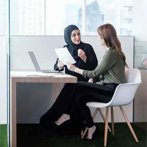 Two professional women, one in abaya and one in business attire, discussing documents in a modern UAE office — symbolising tailored employee benefits advice.